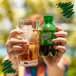 A imagem mostra duas mãos. Uma delas segura uma garrafa pet de 250ml do refrigerante Mate Couro na sua versão tradicional, já a outra mão segura um pequeno copo com a bebida. Ao fundo, a imagem possui um efeito de borrão, por isso não é possível ver o rosto da pessoa. Nos cantos superior direito e inferior esquerdo da imagem, são vistos dois rabiscos que contribuem com a estética da foto.